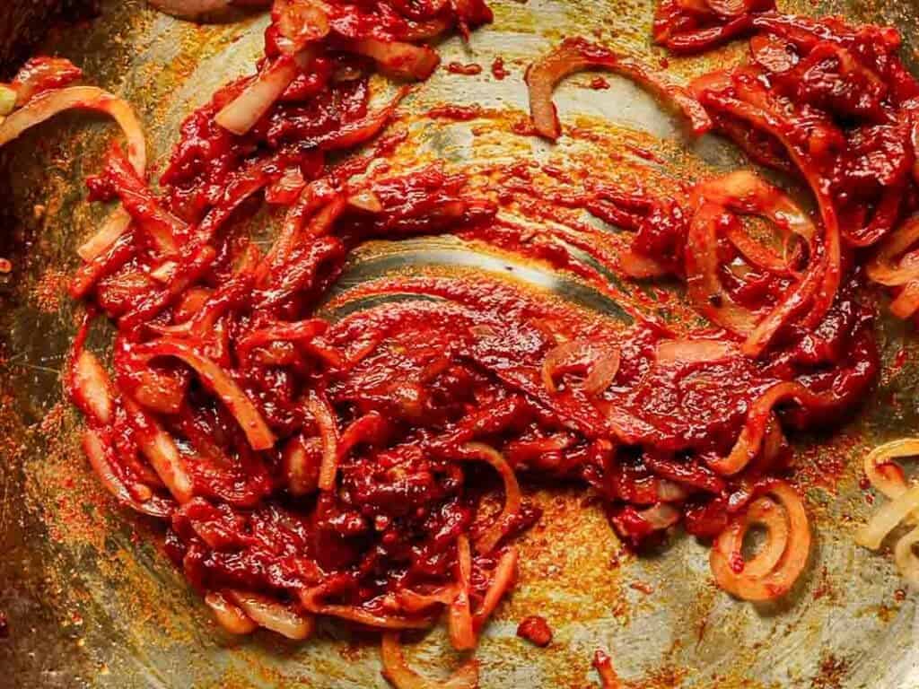 A close-up of vibrant red sauce spread over a textured surface, mixed with sliced shallots. The sauce appears thick and rich, with visible spices and a slightly glossy texture.